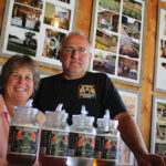 Bill and Lynne Olson along with their lineup of Full Moonshine products. Located behind them are photographs of the Hickory Ledges farm throughout the years.