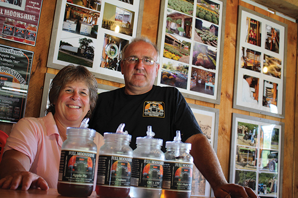 Bill and Lynne Olson along with their lineup of Full Moonshine products. Located behind them are photographs of the Hickory Ledges farm throughout the years.